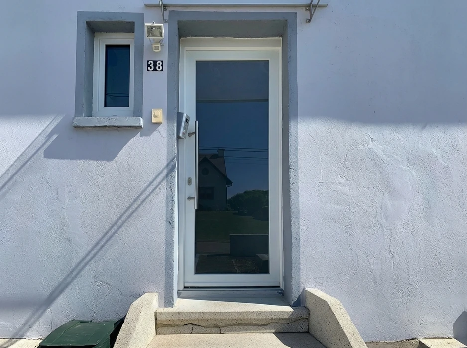 Façade extérieure d'une maison au numéro 38, avec une porte d'entrée blanche à vitrage pleine hauteur fumé, cadre PVC blanc, poignée en aluminium brossé, interphone vidéo encastré