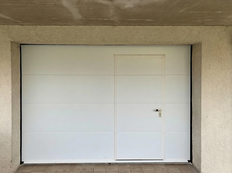 Porte de garage sectionnelle blanche avec portillon intégré, installée dans une baie maçonnée à embrasure en béton texturé